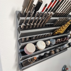 May include: A grey, multi-tiered tool organiser mounted on a white wall, filled with various metal tools. The tools include sockets, screwdriver bits, and grinding stones. The organiser is designed to hold tools of different sizes and shapes.