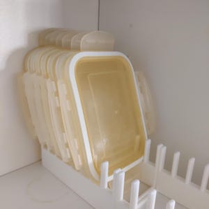Puede incluir: Una pila de recipientes transparentes rectangulares para guardar alimentos con bordes y tapas blancas, cuidadosamente organizados en un organizador de plástico blanco. Los recipientes están dentro de un armario blanco.