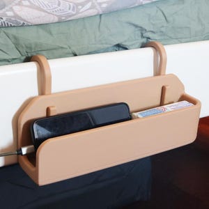 May include: A tan plastic bedside caddy with two curved hooks for mounting onto a bed frame. The rectangular storage container holds a black smartphone connected to a charging cable and a small rectangular package with white and blue text.