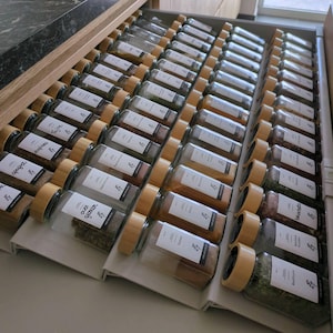 Puede incluir: Un cajón de cocina abierto lleno de frascos de especias de vidrio con tapas de madera, ordenados en filas. Cada frasco tiene una etiqueta blanca con texto. El cajón está en un mueble de madera.