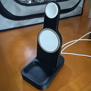 May include: A black charging stand with a white charging pad for a smartwatch and a white charging pad for wireless charging. The stand has a rectangular base and a vertical support with a curved top. A white charging cable is connected.