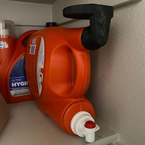 Può includere: Una bottiglia di detersivo arancione con un dispenser nero attaccato a uno scaffale. La bottiglia ha un beccuccio bianco e rosso. Un'altra bottiglia di detersivo è parzialmente visibile sullo sfondo.