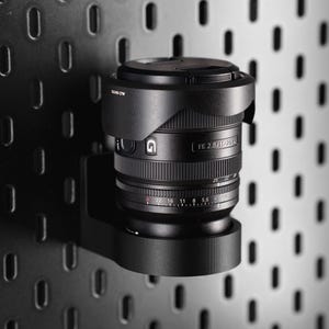 May include: A black camera lens with a lens hood, mounted on a black pegboard. The lens features rings and markings, including aperture settings. The background is a dark grey pegboard.