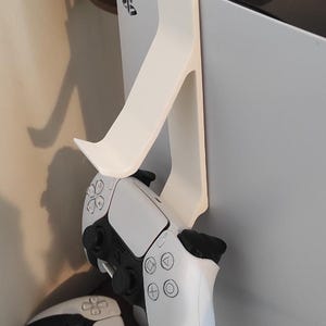 May include: White plastic controller holder for a gaming console, with a white and black controller attached. The holder is mounted on the side of the console. Another controller is visible below.