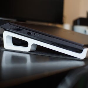 May include: A black laptop computer is elevated on a white laptop stand. The stand is made of a curved, minimalist design. The laptop is positioned at an angle, suggesting improved ergonomics and airflow for the device.