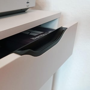 Peut inclure: Un tiroir gris clair avec une poignée noire est partiellement ouvert, révélant un objet noir avec le texte "Bambu Lab". Le tiroir fait partie d'un meuble ou d'un bureau blanc, avec un autre objet blanc visible au-dessus.