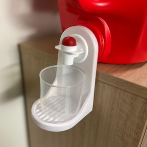 May include: A red plastic container with a white dispenser and a clear measuring cup. The dispenser has a red knob and is attached to a wooden surface. The measuring cup is clear and has measurement markings.