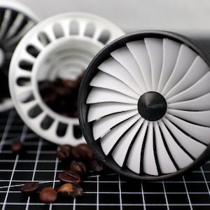 Könnte beinhalten: Drei Kaffee-Artikel, die Turbinen von Strahltriebwerken ähneln. Einer ist schwarz, einer weiß und der dritte weiß mit Kaffeebohnen im Inneren. Die Artikel befinden sich auf einer schwarzen Gitteroberfläche mit verstreuten Kaffeebohnen.