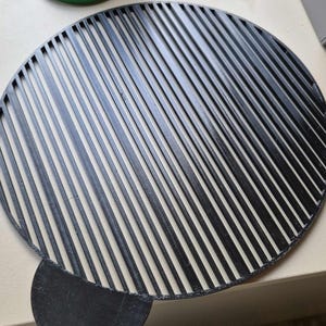 Könnte beinhalten: Ein schwarzer, kreisförmiger Grillrost mit parallelen Linien und einem halbkreisförmigen Griff. Der Rost besteht aus einem dunklen Material und ist zum Grillen von Speisen bestimmt. Das Design ermöglicht eine gleichmäßige Wärmeverteilung.