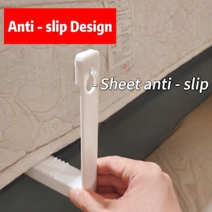 Op de afbeelding: Een wit anti-slip apparaat voor lakens. Het apparaat wordt in de buurt van een matras gehouden. De tekst op de afbeelding luidt "Anti-slip Design" en "Sheet anti-slip".