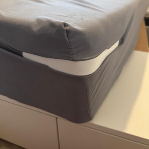 May include: Close-up of a bed with a gray fitted sheet and a white mattress. The sheet is neatly tucked around the mattress, showing a clean, minimalist aesthetic. The bed is on a white platform.