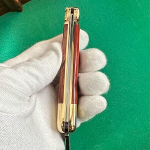 May include: A closed pocket knife with a wooden handle and a gold-colored bolster. The handle is a reddish-brown color with a brass-colored strip down the center. The blade is partially visible and made of steel.