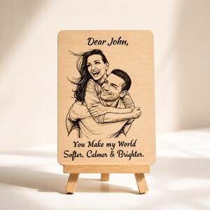 Op de afbeelding: Houten plaat met een zwart-wit illustratie van een omhelzend stel. De tekst "Dear John, You Make my World Softer, Calmer & Brighter" is op de plaat gedrukt. De plaat wordt gepresenteerd op een kleine houten ezel.