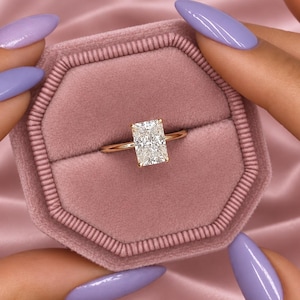 Op de afbeelding: Een gouden ring met een enkele rechthoekige, heldere edelsteen in radiant-slijpvorm, gezet in een zetting met vier pootjes. De ring wordt gepresenteerd in een achthoekig, oudroze fluwelen doosje met een gleuf in het midden.