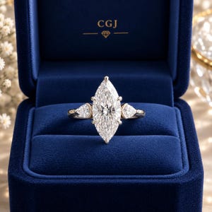 May include: A marquise-cut diamond ring featuring a large central stone flanked by two smaller pear-shaped diamonds on a polished metal band, presented inside a deep blue velvet box with the gold text CGJ and a diamond icon printed inside.