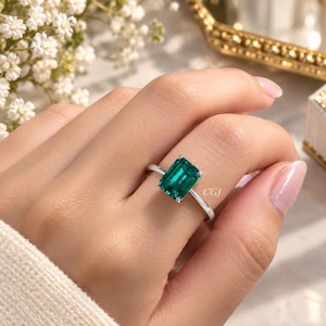 Peut inclure: Une bague argentée ornée d'une grande pierre précieuse verte de taille émeraude rectangulaire, fixée par quatre griffes. L'anneau est fin et poli, avec les lettres CGJ discrètement gravées sur le côté de la monture métallique près de la pierre.