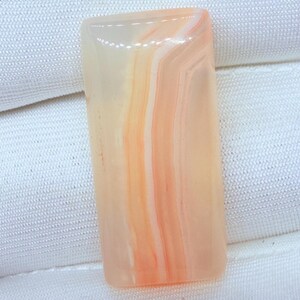 May include: A rectangular cabochon gemstone featuring a smooth, polished surface with translucent layers of pale peach, cream, and soft orange hues. The stone displays natural banding patterns that run vertically along its length.