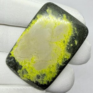 Pode incluir: Um cabochão de pedra preciosa polida de formato retangular, apresentando um centro liso em bege claro rodeado por uma borda texturizada em verde chartreuse vibrante e preto carvão, com acabamento superficial brilhante.