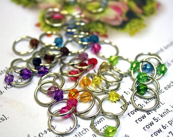 May include: A selection of silver stitch markers, each with a colourful faceted bead in shades of purple, red, orange, yellow, green, and blue. The markers are scattered on a white surface with some text visible. Perfect for knitting and crochet.