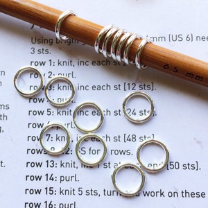 May include: Close-up of silver knitting stitch markers on a wooden knitting needle. The needle is labelled "6.5 mm (US 6)".