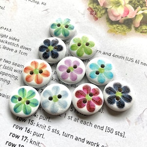 May include: Ten round wooden buttons with a floral design. Each button has a white base and a painted flower in colours like green, blue, purple, orange, and red. The buttons have two holes for sewing. The buttons are approximately 2.5 cm in diameter.