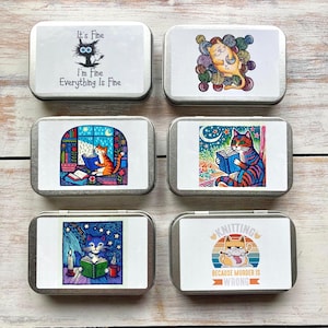 May include: Six small, rectangular metal tins with cat-themed illustrations. One tin reads "It's Fine, I'm Fine, Everything Is Fine." Other tins feature cats with yarn, reading books, and the text "Knitting Because Murder Is Wrong."