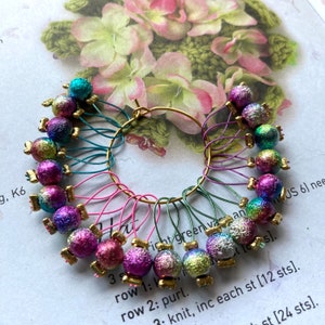 May include: A set of 18 colourful stitch markers with iridescent glitter, arranged in a circle on a gold ring. The markers are attached to the ring with different coloured cords.