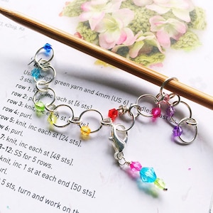 以下が含まれることがあります： 虹色のビーズが付いた銀色の留め具が付いたステッチマーカー。マーカーは木製の編み針に取り付けられています。紙のテキストは次のとおりです。「明るい緑色の糸と 4mm（US 6）の針を 3 目の後に使用します。1 段目：k、各目ごとに増し目 [6 目] 2 段目：purl 3 段目：k、各目ごとに増し目 [24 目]。 」