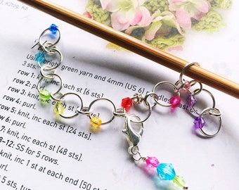 May include: A rainbow colored beaded stitch marker with a silver clasp. The marker is attached to a wooden knitting needle. The text on the paper reads: "Use bright green yarn and 4mm (US 6) needles past 3 sts. row 1: k, inc each st [6 sts] row 2: purl row 3: knit, inc each st [24 sts]."