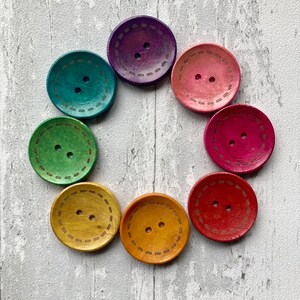 May include: Seven wooden buttons in various colours, including purple, pink, red, green, and yellow. Each button has two holes and a decorative stitched design around the edge.