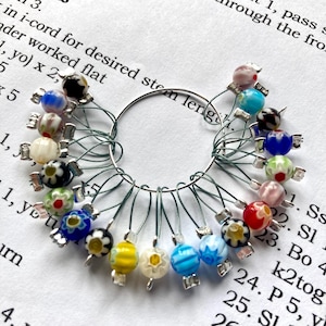 May include: A selection of colourful glass bead stitch markers attached to a silver ring. The beads are various colours, including blue, red, yellow, and white, with floral and abstract designs. The markers are on a white surface with text.