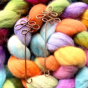 May include: Two copper wire knitting needle holders with a decorative knot design. The holders are displayed against a background of colourful yarn.