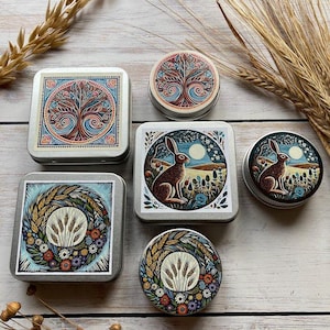 May include: A selection of decorative metal tins with vibrant illustrations. The tins showcase designs including a tree of life, a rabbit in a field under a full moon, and wheat encircled by flowers. The tins are square and circular.