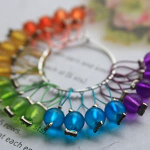 May include: A set of colorful stitch markers for knitting or crocheting. The markers are made of glass beads and metal rings. The beads are arranged in a rainbow pattern, with orange, yellow, green, blue, and purple beads. The markers are a useful tool for keeping track of rows and stitches in knitting and crocheting projects.
