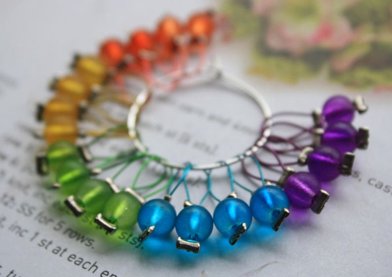 May include: A set of colorful stitch markers for knitting or crocheting. The markers are made of glass beads and metal rings. The beads are arranged in a rainbow pattern, with orange, yellow, green, blue, and purple beads. The markers are a useful tool for keeping track of rows and stitches in knitting and crocheting projects.