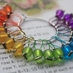 May include: A rainbow of colorful glass beads on a silver ring, used as stitch markers for knitting or crocheting. The beads are arranged in a circle, with red, orange, yellow, green, blue, and purple beads.