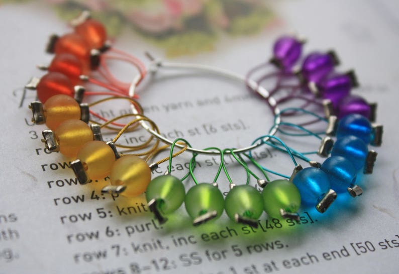 May include: A rainbow of colorful glass beads on a silver ring, used as stitch markers for knitting or crocheting. The beads are arranged in a circle, with red, orange, yellow, green, blue, and purple beads.