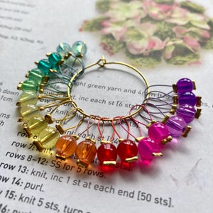 May include: A collection of rainbow-coloured stitch markers for knitting or crochet. Each marker has a different coloured bead, attached to a gold-coloured ring. The markers are designed to help track stitches in a knitting or crochet project.