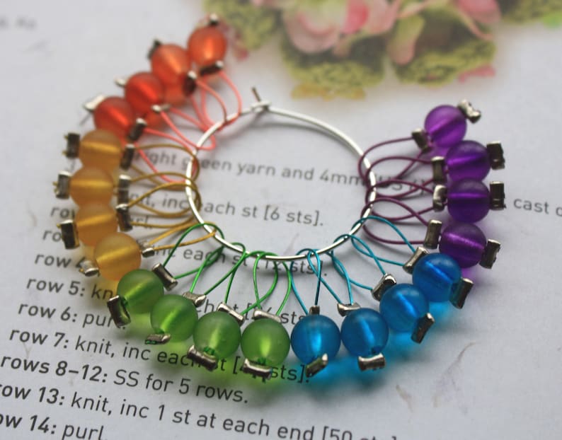 May include: A set of colorful stitch markers in a rainbow pattern. The markers are made of glass beads and metal rings. The beads are in shades of orange, yellow, green, blue, and purple. The markers are attached to a silver ring.