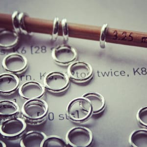 May include: A close-up of a pile of silver jump rings, with a wooden knitting needle in the background. The needle is labelled "3.25 mm".