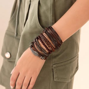 May include: A multi-layered bracelet set featuring dark brown leather cords, a braided leather strand, a row of dark red beads, and a metallic charm shaped like a wing. The set consists of several individual bands worn together on the wrist.
