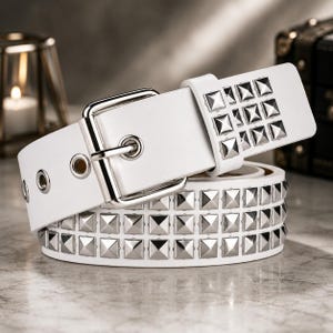 May include: A white belt featuring two rows of silver-toned pyramid-shaped metal studs and a silver-toned metal buckle. The belt includes a matching white keeper loop and a series of silver-toned metal grommets along the strap length.