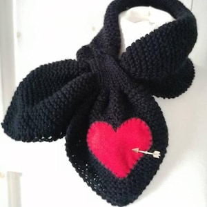 May include: A black knitted scarf featuring a red heart and a gold-coloured arrow pin. The scarf is made from a thick, textured yarn. The heart is appliquéd onto the scarf. A winter accessory, suitable for Valentine's Day or a romantic occasion.