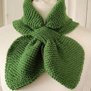 May include: A green knitted scarf displayed on a white mannequin. The scarf is a vibrant shade of green, with a textured, hand-knitted appearance. The scarf is designed to be worn around the neck, with a unique, folded design.