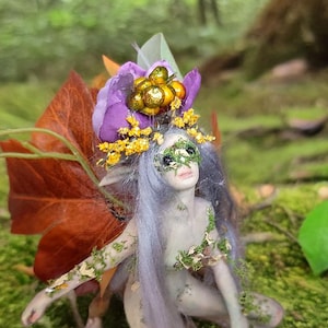 Op de afbeelding: Een miniatuur feeënbeeldje met een paarse en gouden hoofdtooi, groen masker en bladachtige vleugels. De fee knielt op een bed van groen mos, omringd door gebladerte.