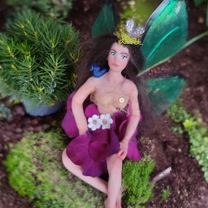 Puede incluir: Una figura de hada hecha a mano con una falda de pétalos morados y alas verdes y negras. El hada tiene cabello castaño, ojos azules y un pequeño pájaro azul en el hombro. Lleva una parte superior dorada y una corona plateada con detalles amarillos. El hada está rodeada de vegetación.