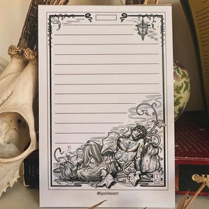 May include: A lined notepad featuring a black and white illustration of a reclining figure with tattoos, surrounded by leaves and pumpkins. The notepad has a decorative border and the text "@igildasart".