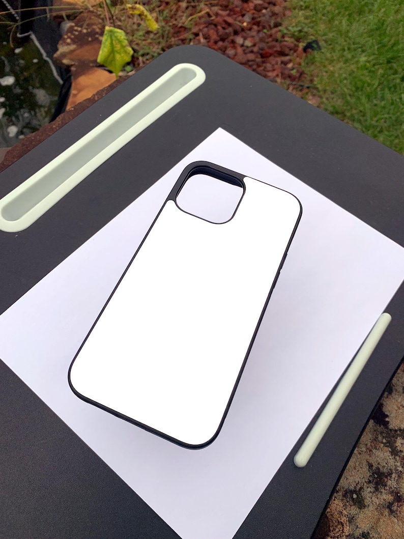 Puede incluir: Una funda de tel&eacute;fono blanca con borde negro, sobre una hoja de papel blanca. Un l&aacute;piz &oacute;ptico verde claro y un escritorio negro con una ranura verde claro tambi&eacute;n son visibles. La funda est&aacute; en blanco.