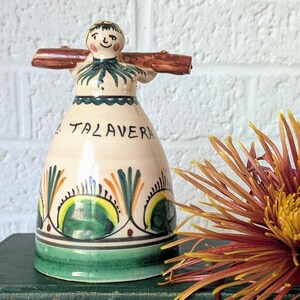 May include: A ceramic bell-shaped figurine with a painted face and wings, labeled "Talavera." It sits atop a stack of vintage books, with a large, spiky orange and yellow flower to the right. The books are green and the background is a white brick wall.