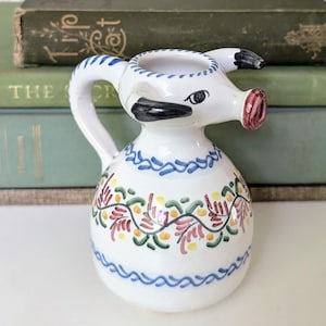 May include: A white ceramic pitcher with a pig-like design, featuring a handle and a spout. The pitcher is decorated with blue, pink, and green floral patterns. It is placed in front of a stack of green books and a potted plant.